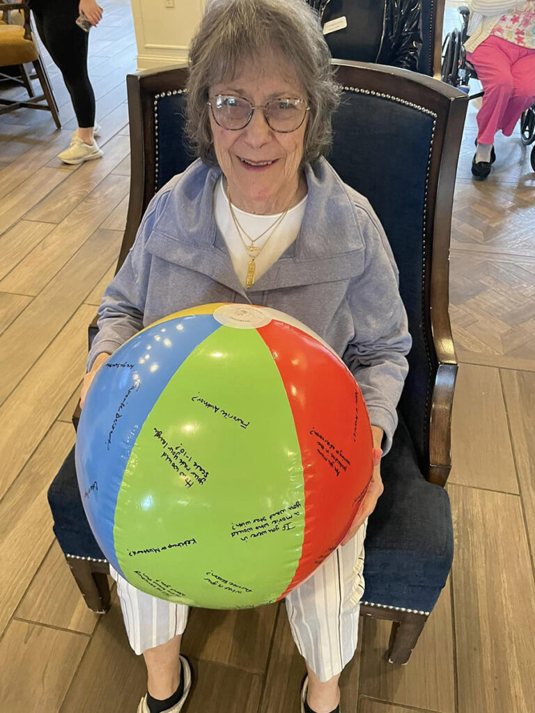 With a joyful smile, an older woman sits in a chair, gripping a colorful beach ball, during an Employee Orientation for Beachball chair volleyball session.