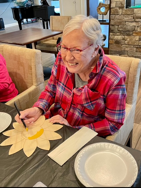 An elderly woman happily painting a flower, part of a fun activity with other senior residents.