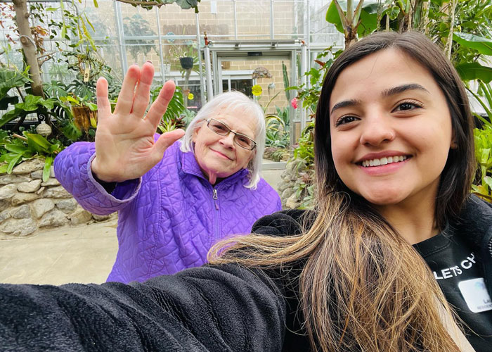 A senior resident in a purple jacket waves while taking a selfie with a younger woman, smiling together in a lush greenhouse, capturing a joyful moment of companionship.