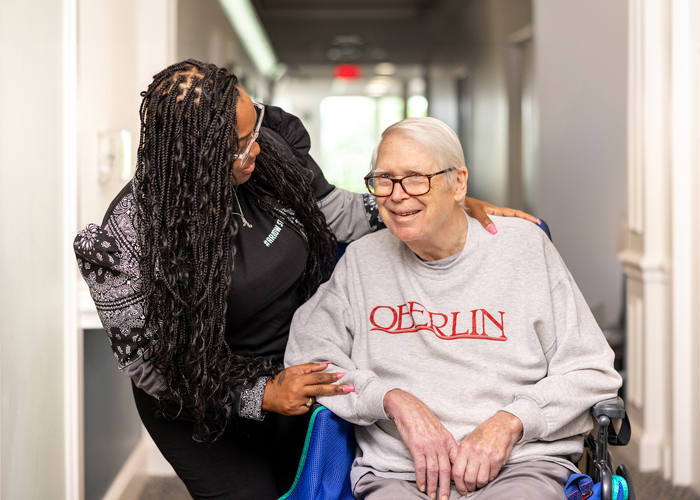 Friendly caregiver smiles and chats with a senior resident in a wheelchair, who is wearing an Oberlin sweatshirt, showcasing compassion and connection in a senior living community.