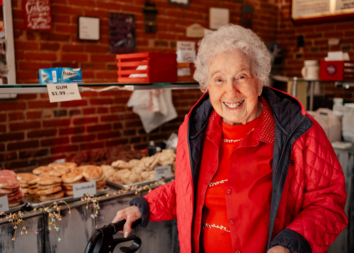 Senior resident in a red jacket smiles while standing with a walker in front of a bakery counter displaying cookies and treats inside a cozy shop with brick walls.