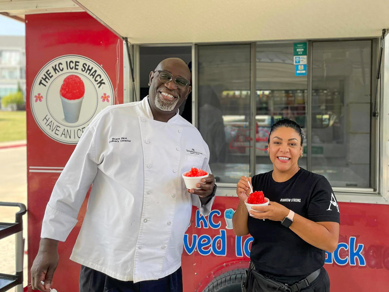 A senior living community's Culinary Director and a staff member smile while holding bowls of shaved ice in front of a shaved ice truck during a summer event.