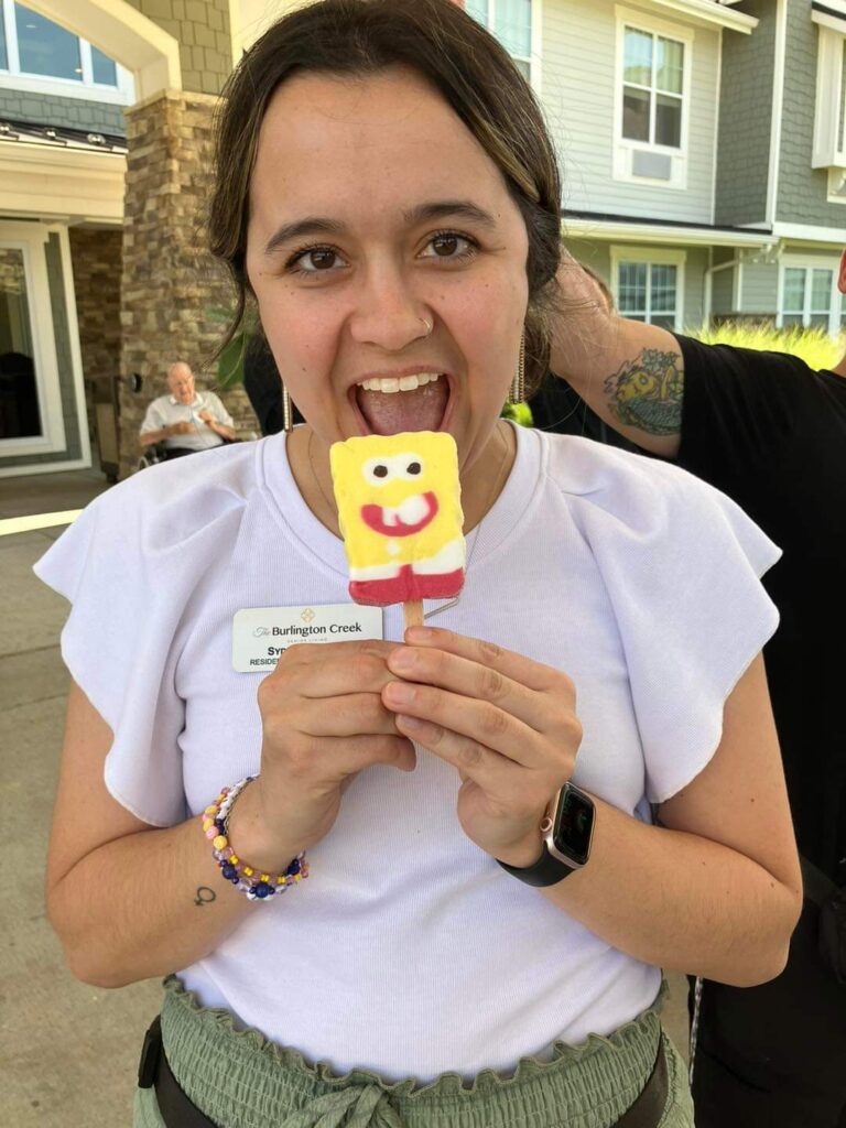 Employee smiling and holding a character-shaped ice cream treat outside a senior living community.