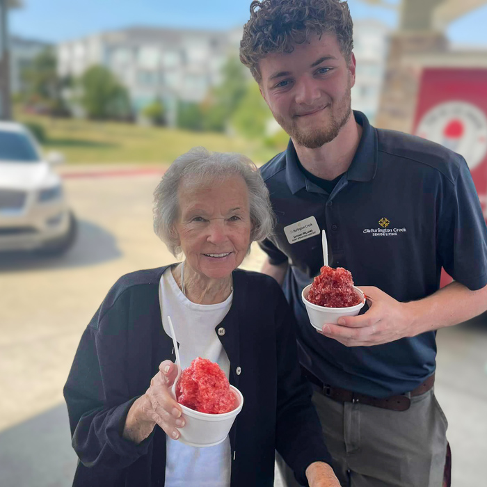 A senior resident and a staff member hold shaved ice bowls, smiling happily outdoors at a summer event hosted at a senior living community.