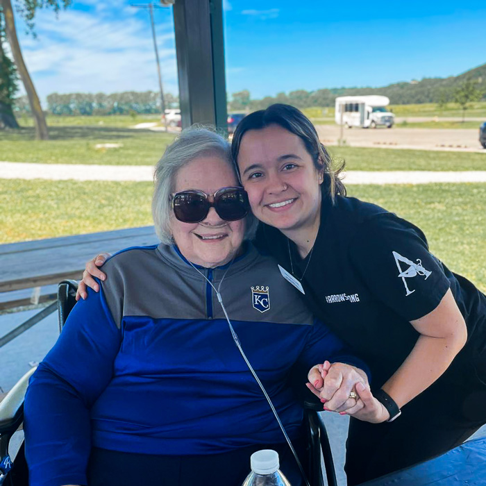 A senior resident wearing sunglasses sits beside a staff member, both smiling and enjoying time outdoors during a sunny day at a senior community park event.